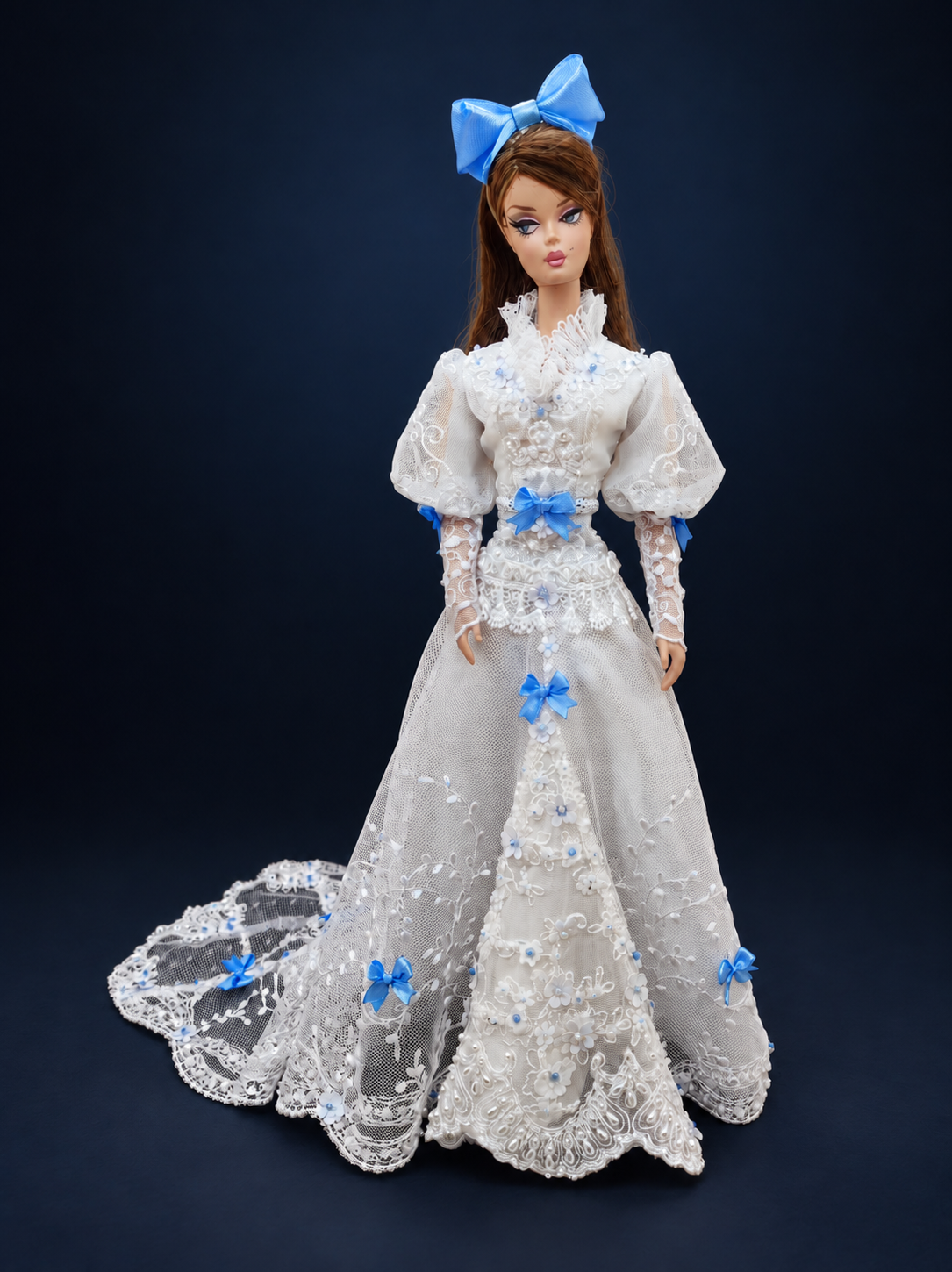 Three-quarter front view of a fashion doll wearing a white lace gown with blue bows, puff sleeves and a long embroidered train. On a dark navy studio background.
