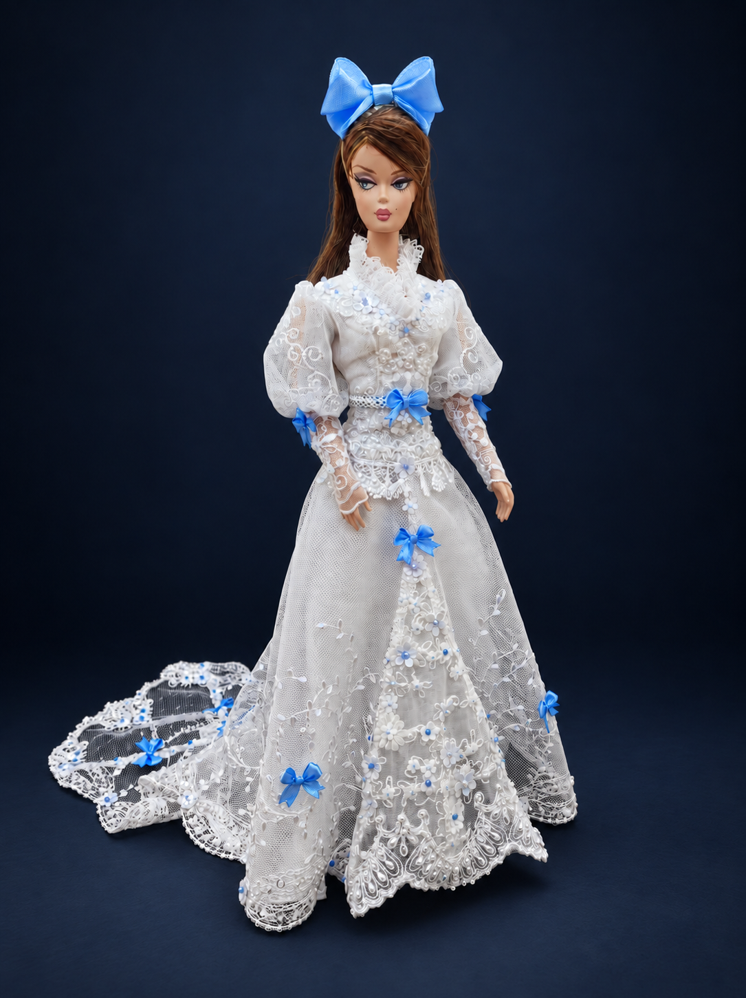 Front view of a white lace fashion doll gown with a high collar, blue satin bows, floral embroidery and a matching bow headpiece. On a dark navy studio background.