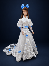 Front view of a white lace fashion doll gown with a high collar, blue satin bows, floral embroidery and a matching bow headpiece. On a dark navy studio background.