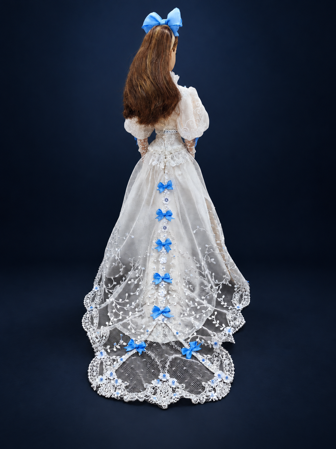 Back view of a white lace fashion doll gown with a row of blue bows and a sheer embroidered train. On a dark navy studio background.