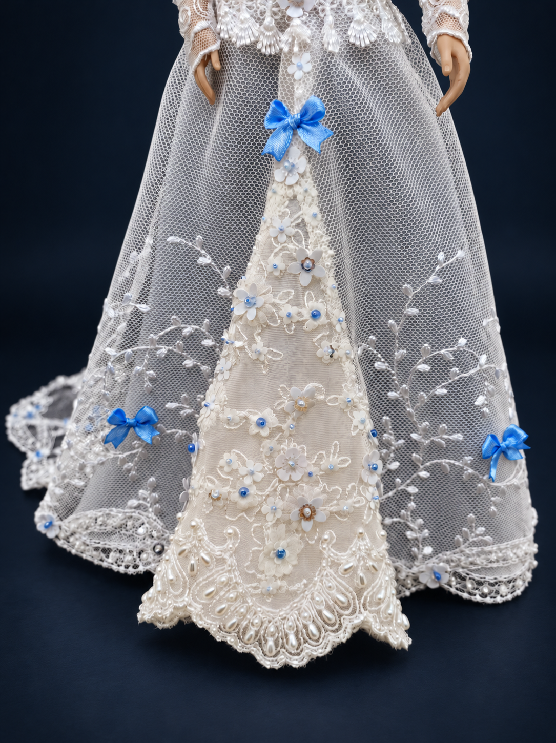 Close-up of the white lace doll gown skirt, showing blue bows, floral appliques, beads and embroidered scalloped edging. On a dark navy studio background.