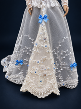 Close-up of the white lace doll gown skirt, showing blue bows, floral appliques, beads and embroidered scalloped edging. On a dark navy studio background.