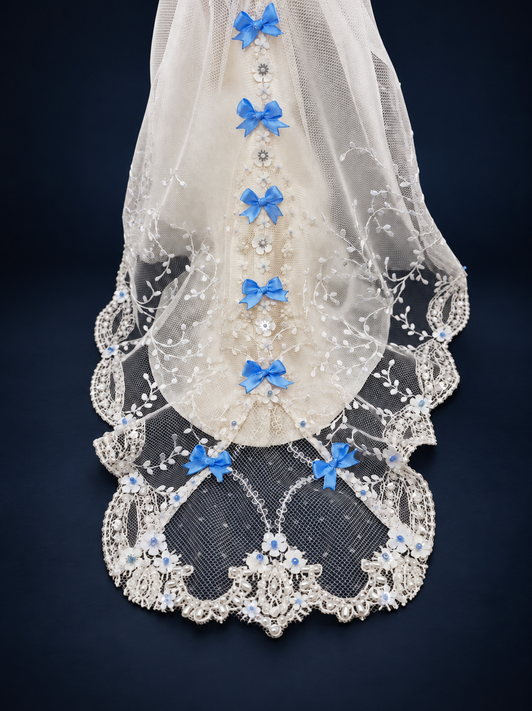 Close-up of the doll gown's sheer lace train with blue satin bows, white floral appliques and decorative beaded edging. On a dark navy studio background.