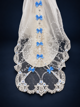 Close-up of the doll gown's sheer lace train with blue satin bows, white floral appliques and decorative beaded edging. On a dark navy studio background.