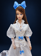 Close-up of the doll's blue bow headpiece and white lace gown bodice with a high collar, puff sleeves and floral embellishments. On a dark navy studio background.