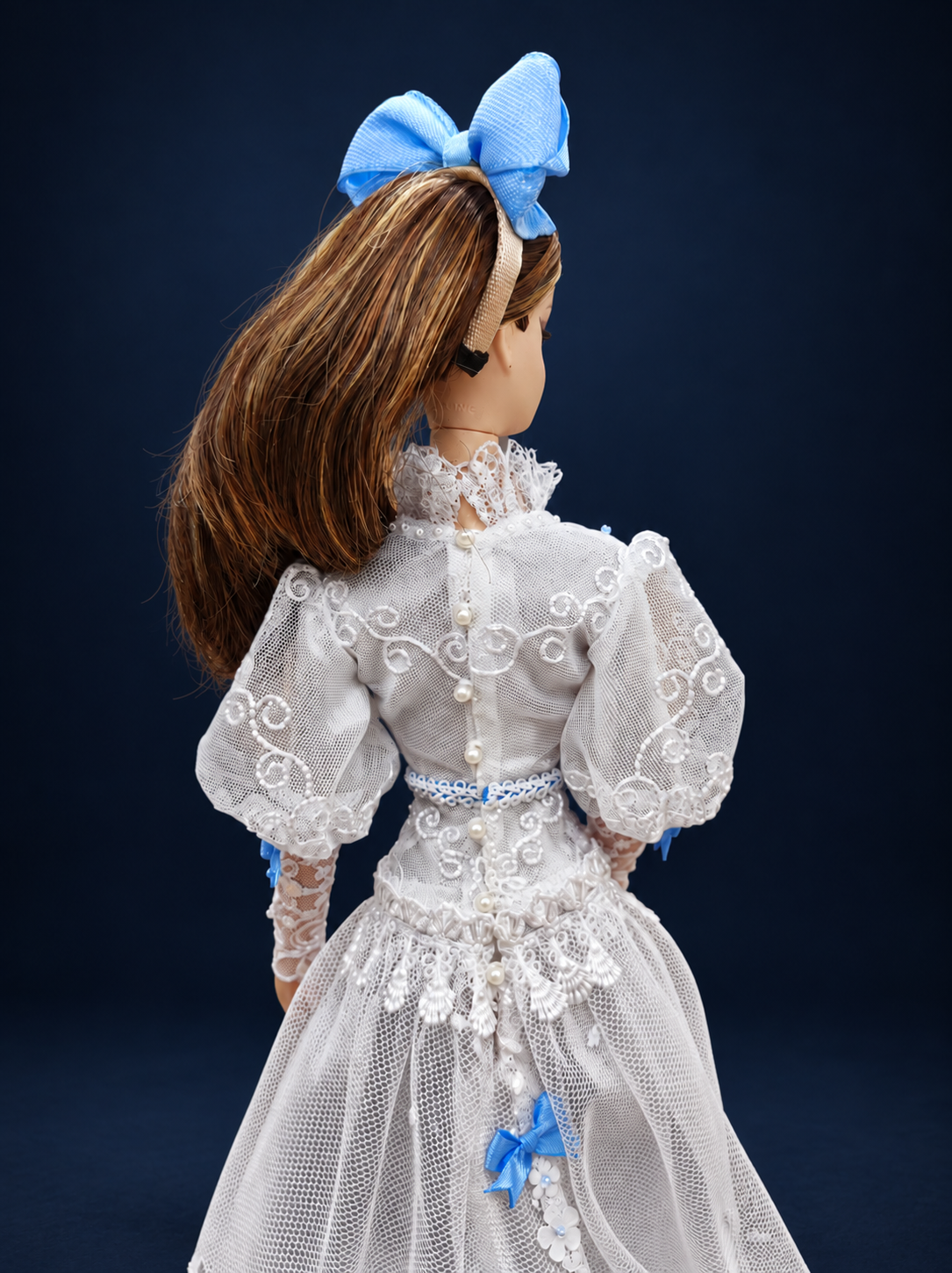 Rear close-up of the white lace doll gown showing pearl-like buttons, embroidered puff sleeves, a blue waist ribbon and lace cuffs. On a dark navy studio background.