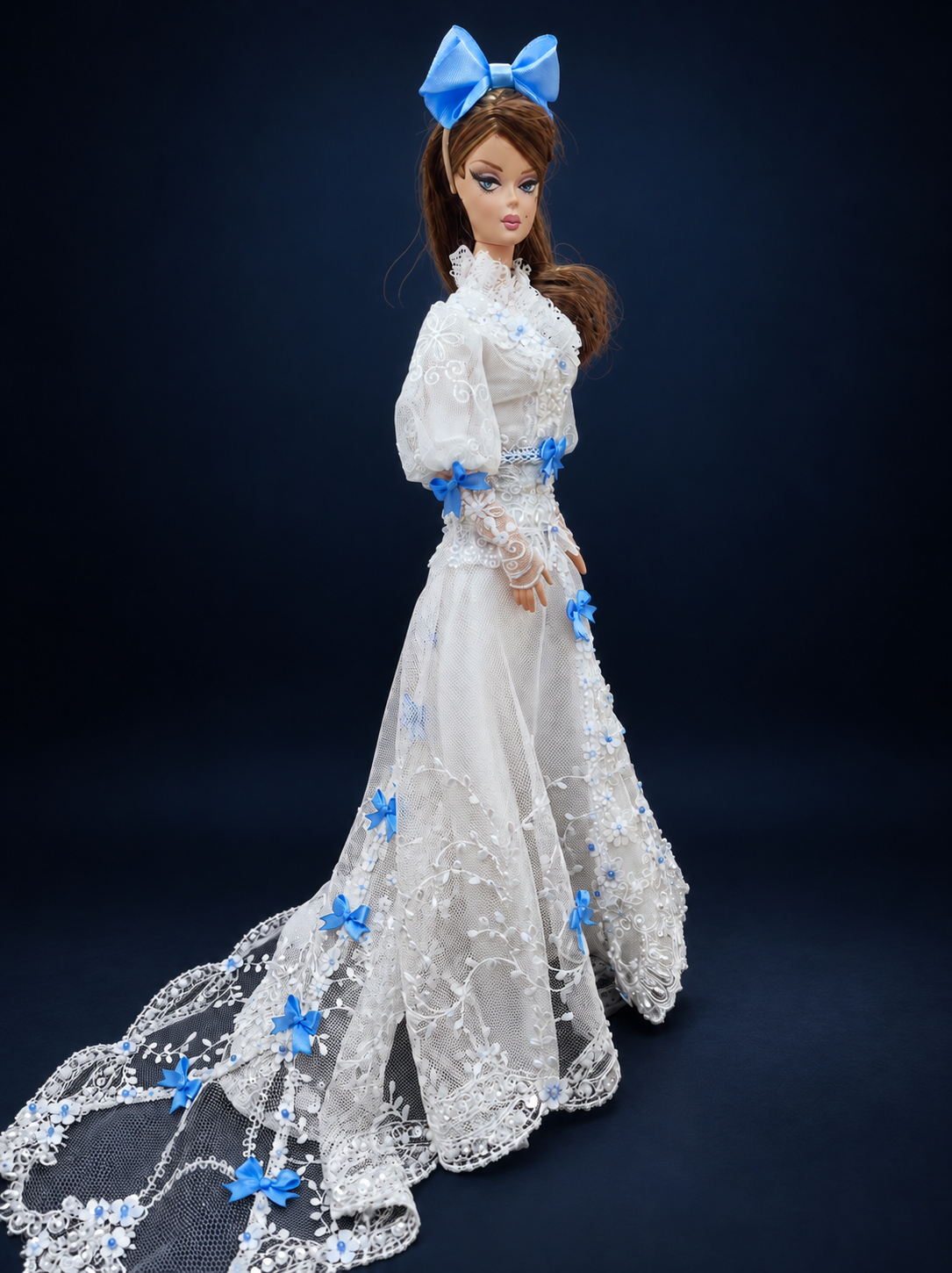 Side view of a fashion doll in a white lace gown with blue bows and a long sheer train extending to the left. On a dark navy studio background.