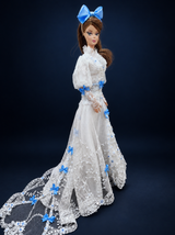 Side view of a fashion doll in a white lace gown with blue bows and a long sheer train extending to the left. On a dark navy studio background.