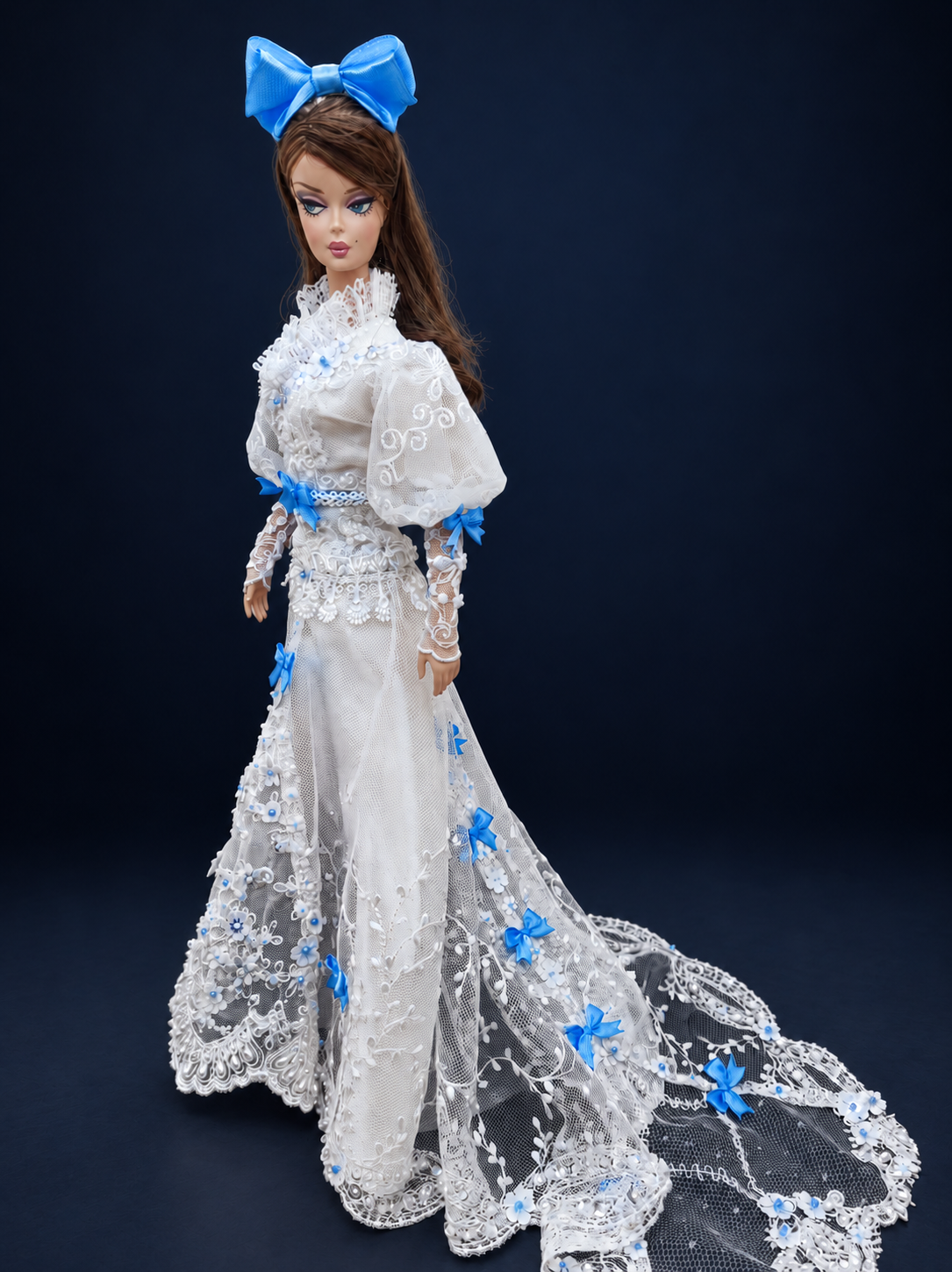 Side view of a fashion doll in a white lace gown with blue bows, showing floral embroidery and a sheer train extending to the right. On a dark navy studio background.